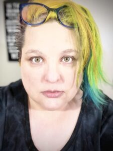 Accidental selfie of Raven Oak featuring blue computer glasses, rainbow hair, gray shirt, and a wide-eyed stare.