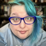 Author & artist Raven Oak with green eyes, blue/teal hair (with a side shave), blue shirt, and blue glasses. They are sitting in front of their office library.