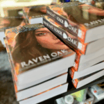 Two stacks of books are sitting on the table. The books are copies of Amaskan's Honor by Raven Oak and are thick paperbacks.