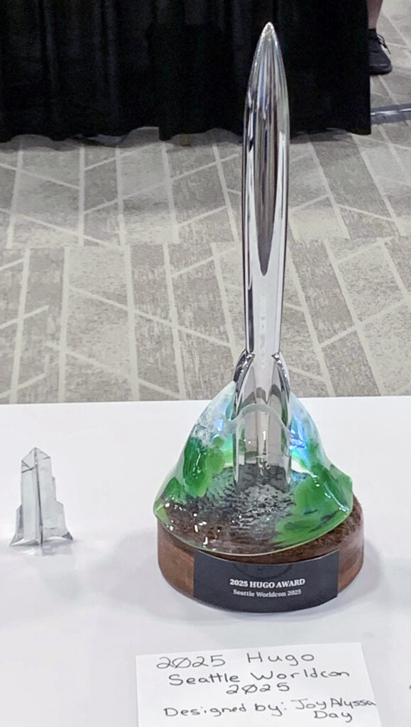 A photo of the 2025 Hugo Award featuring a rocket coming out of a green forest and mountain. Designed by Joy Alyssa Day.