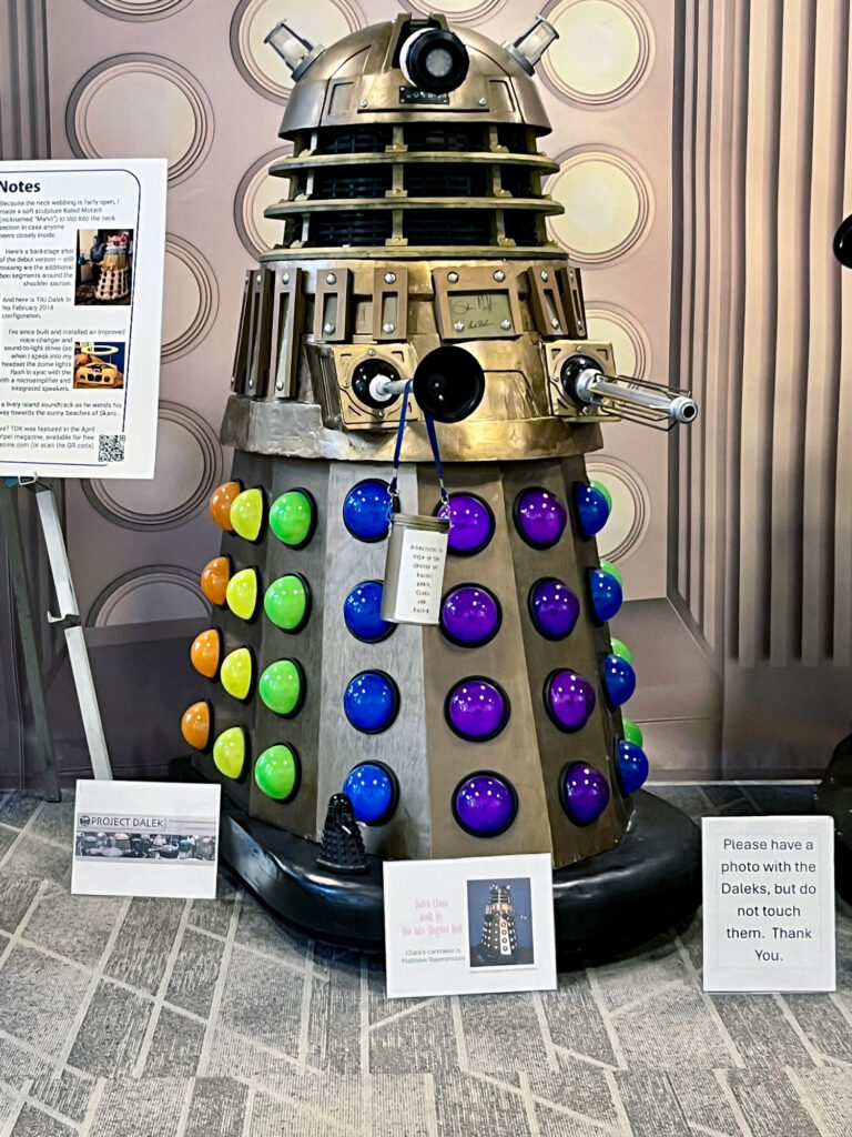 A rainbow colored Dalek named Clara is in the photo. She was built by the late Shaylee Bell.