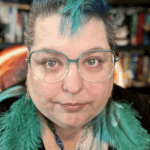 Author & artist Raven Oak wearing no make-up but with a teal faux-mohawk and teal feather earrings on. Teal glasses as well. Photo from 9/1/25.
