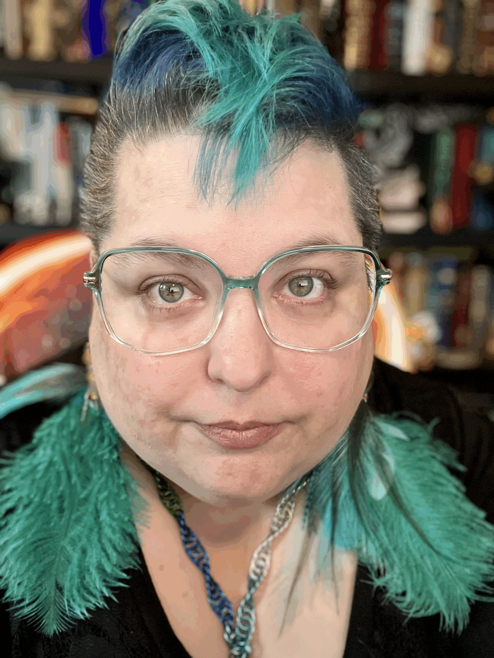 Author & artist Raven Oak wearing no make-up but with a teal faux-mohawk and teal feather earrings on. Teal glasses as well. Photo from 9/1/25.